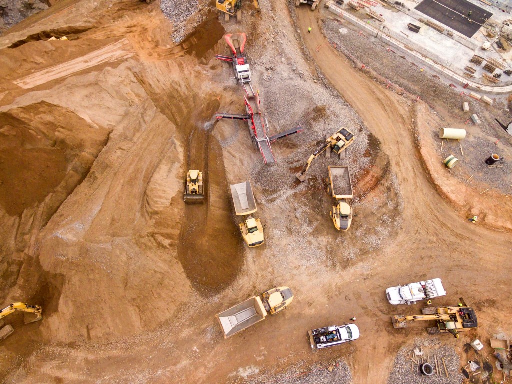 An overhead shot of an active construction site where heavy machinery is engaged in land development. Bulldozers, excavators, and dump trucks are strategically positioned across the terrain, sculpting the earth and laying the groundwork for infrastructure. Amidst piles of dirt and construction materials, the scene captures the early stages of preparing the land for utilities such as water and electricity, all under the supervision of specialized land development services to minimize environmental impact.