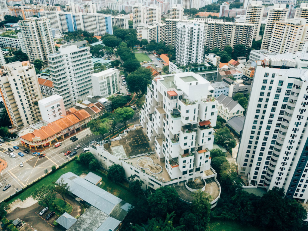 An aerial view of a mixed-use urban development, showcasing a variety of building structures including high-rise residential towers, mid-rise apartments with balconies and rooftop gardens, and traditional low-rise shophouses with red-tiled roofs. The area seamlessly transitions from a dense urban residential zone to a commercial district, illustrating the transformation of raw land to accommodate housing and retail spaces. There's a visible contrast between the old and new architectures, hinting at the evolution of the area's land use.
