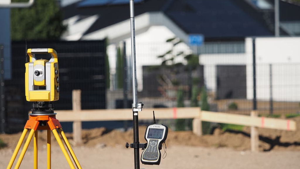 This image displays a surveying equipment setup in a residential construction area. In the foreground, there's a precise yellow theodolite on a tripod, a tool used for measuring horizontal and vertical angles, indicating the process of a boundary survey. Next to it, there's an electronic distance meter (EDM) on a pole, used to measure distances accurately. The blurred background features a residential setting with a modern house and a fence, which could be the subject of the survey. This setting evokes the moment of conducting a boundary survey, where the clarity of property lines is essential.